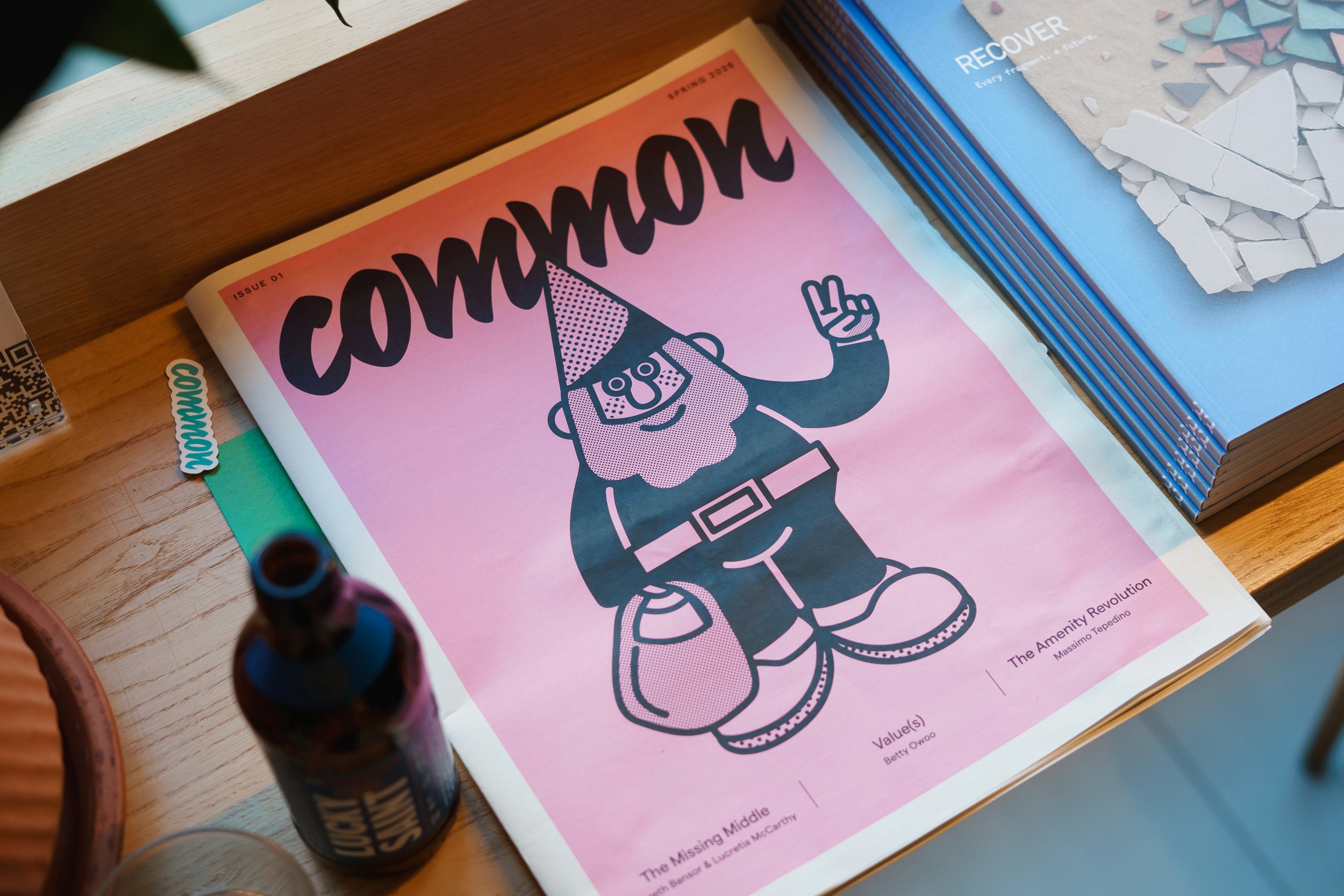 Pink Common magazine cover with a gnome illustration giving a peace sign, on a wooden shelf